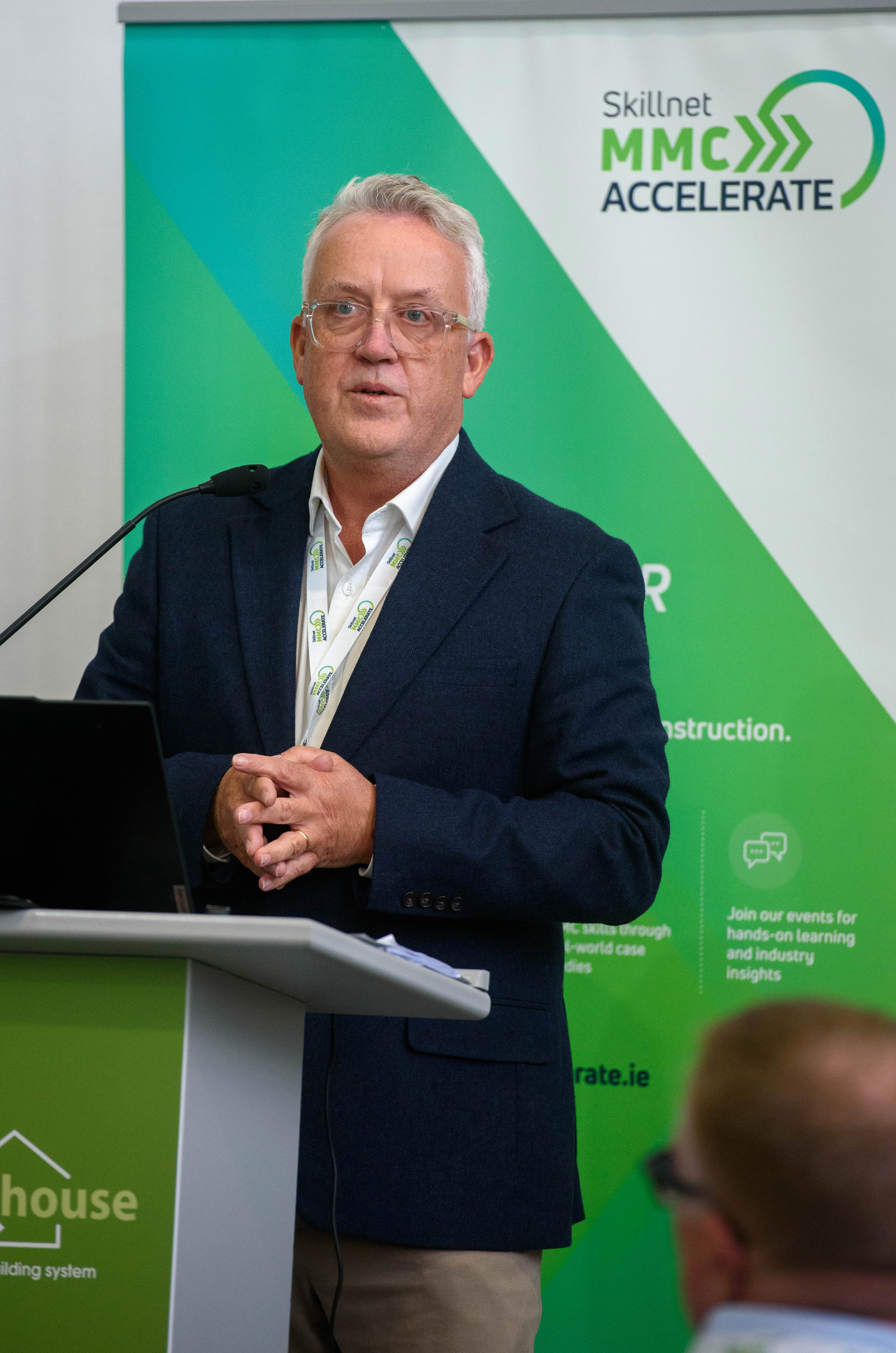 A person in a suit speaking at a podium with a green background featuring a logo.