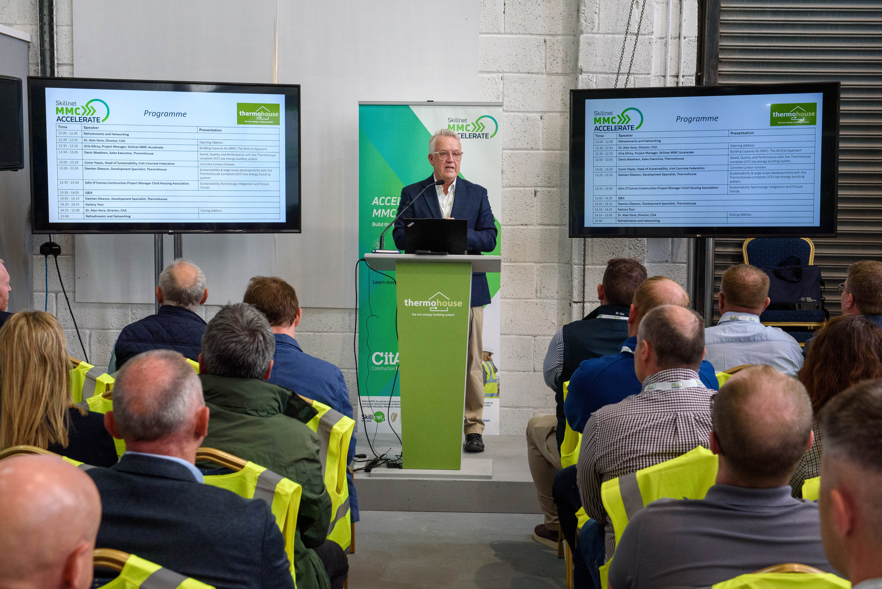 Speaker presenting at a podium with green backdrop to an audience of seated attendees.