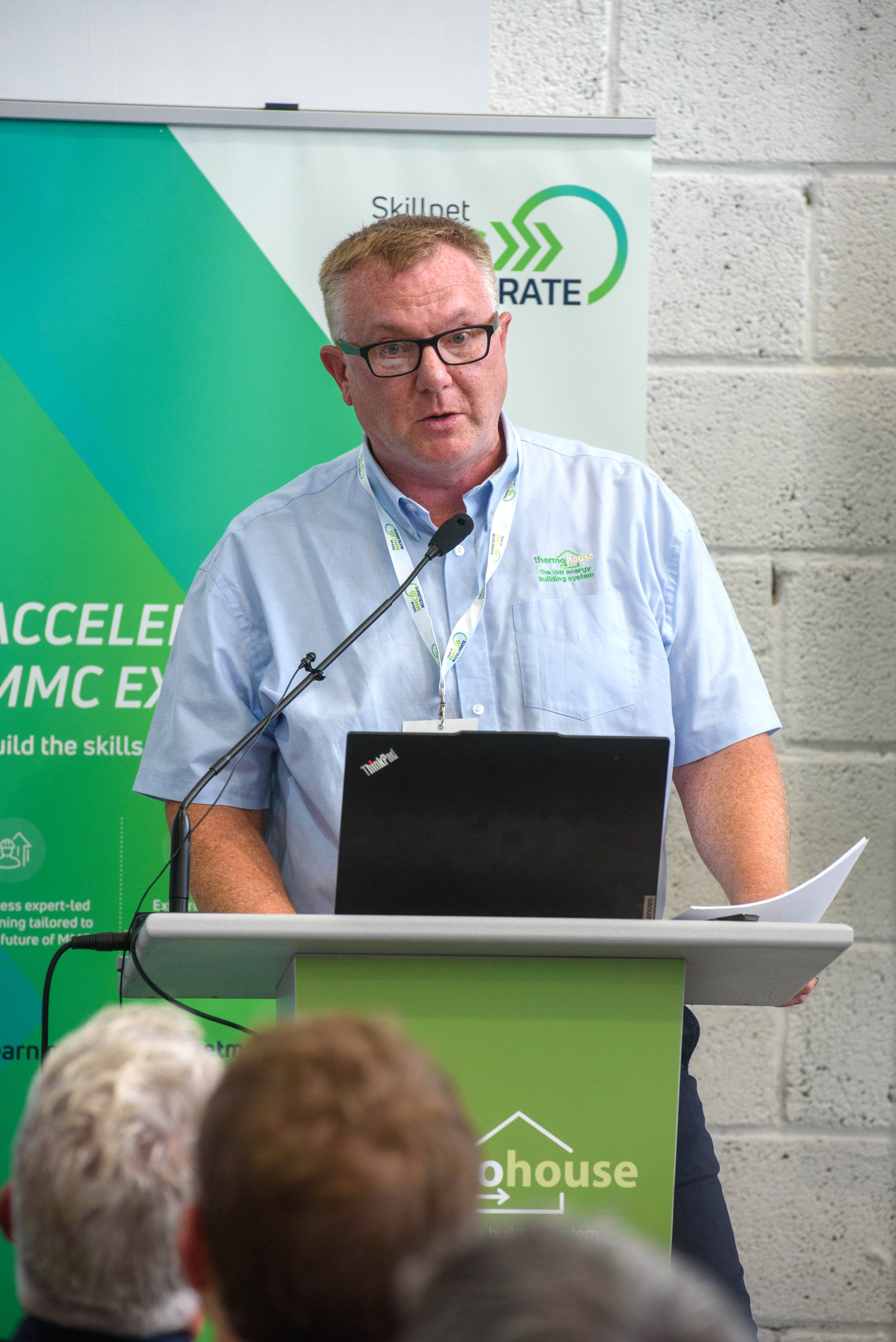 A person in a blue shirt speaking at a podium with a green backdrop and logo.