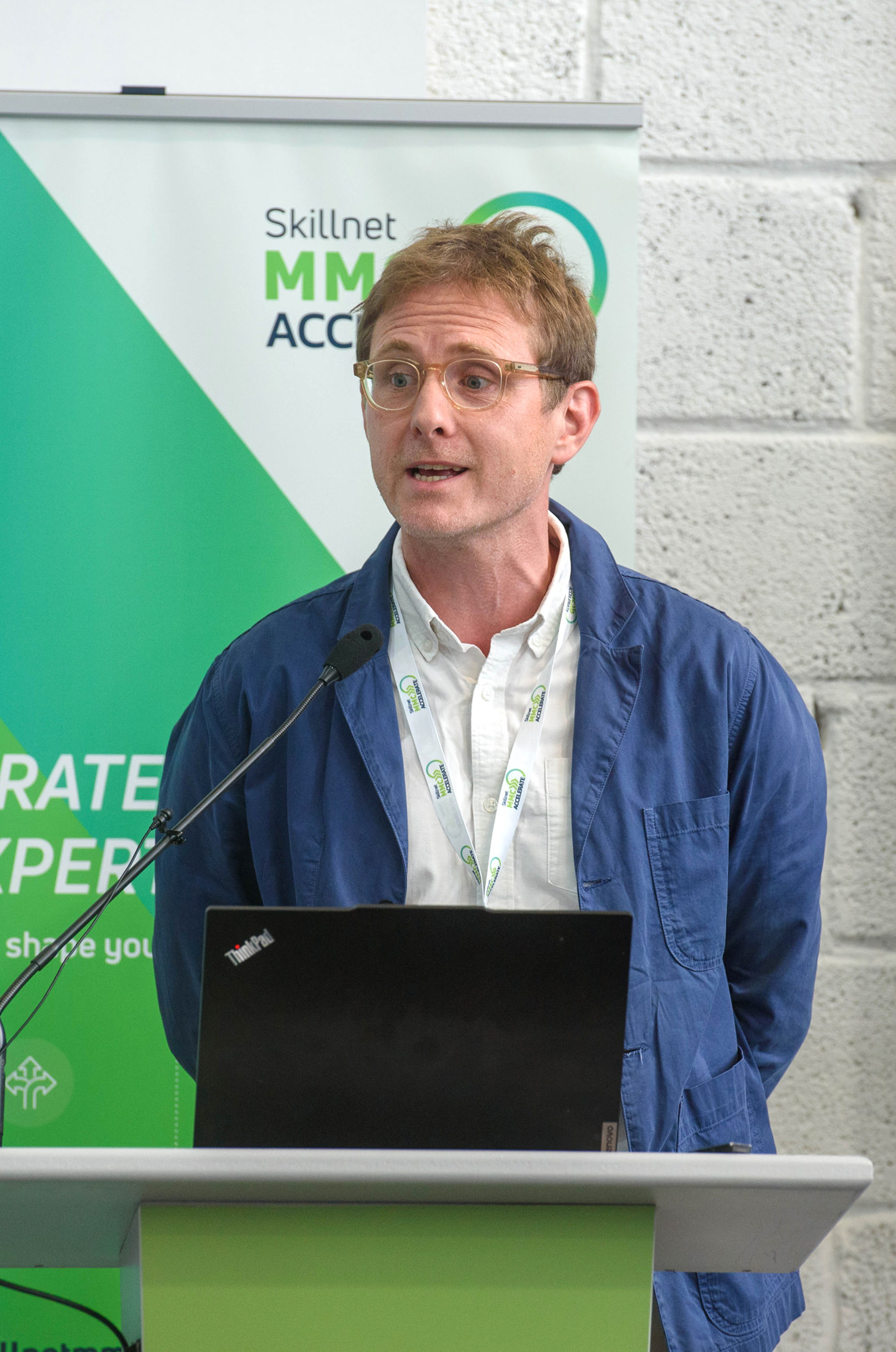 A person in a blue jacket standing at a podium with a laptop and green banner behind them.