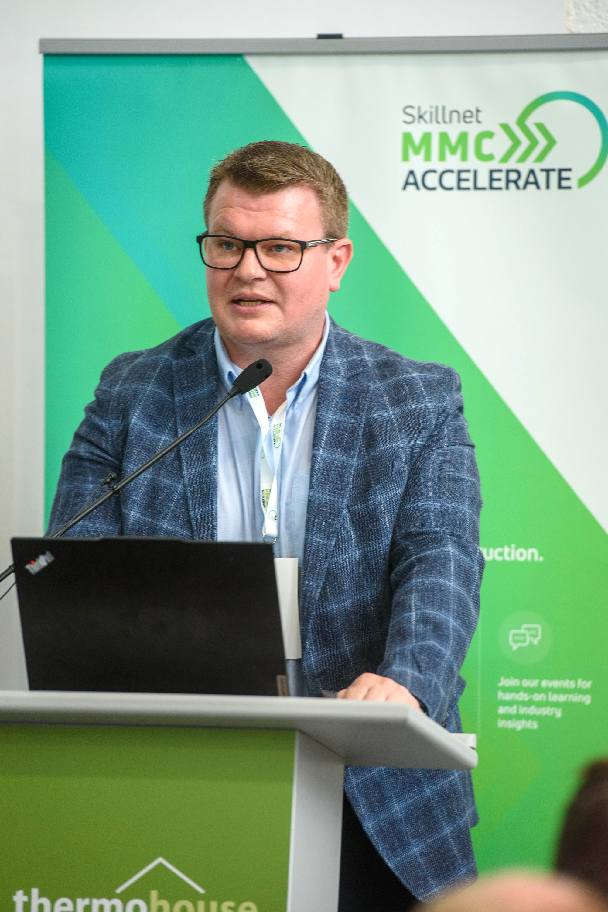 A person in plaid blazer at podium with green APEC backdrop and laptop.