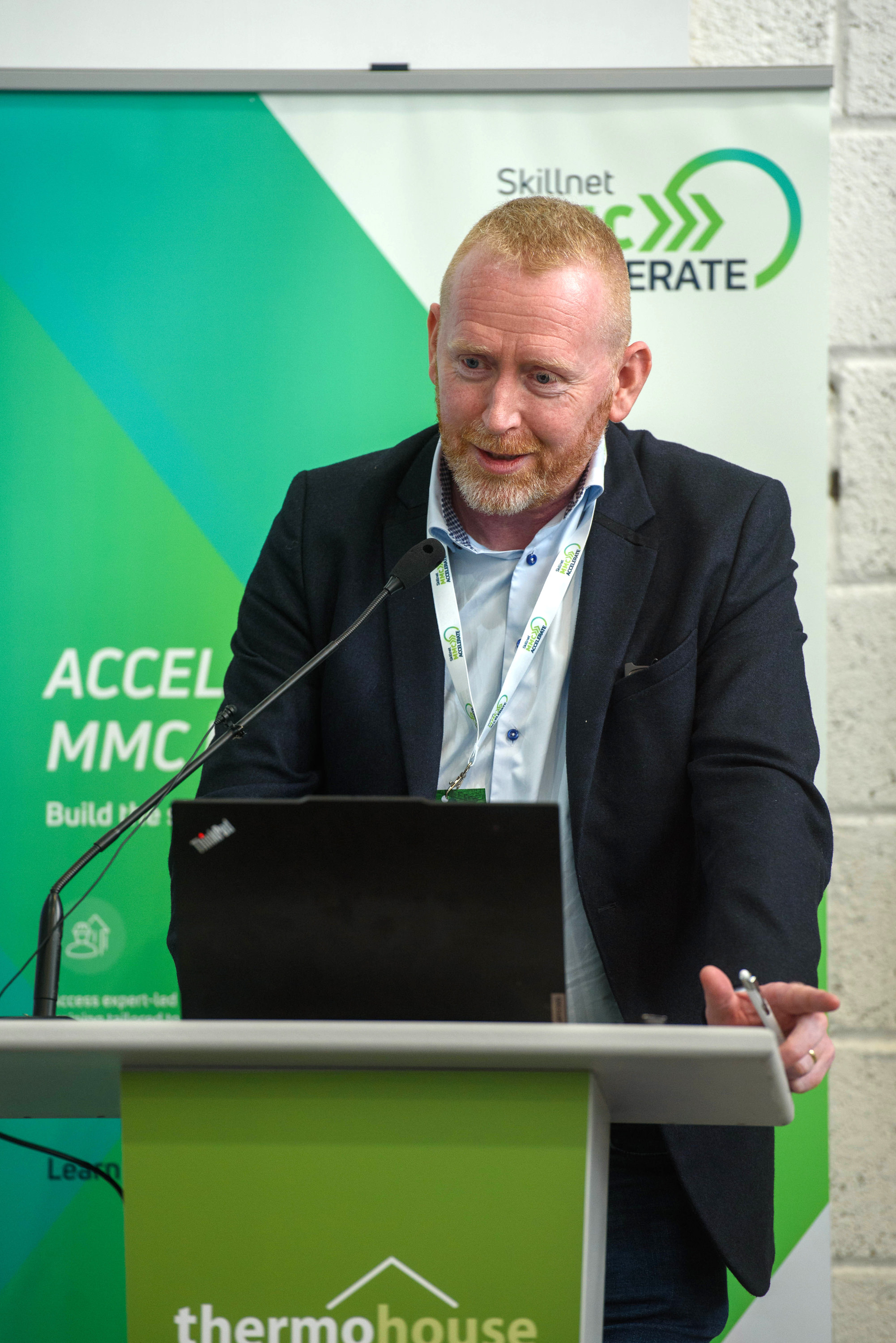 Person in a suit speaking at a podium with a green backdrop featuring a logo.
