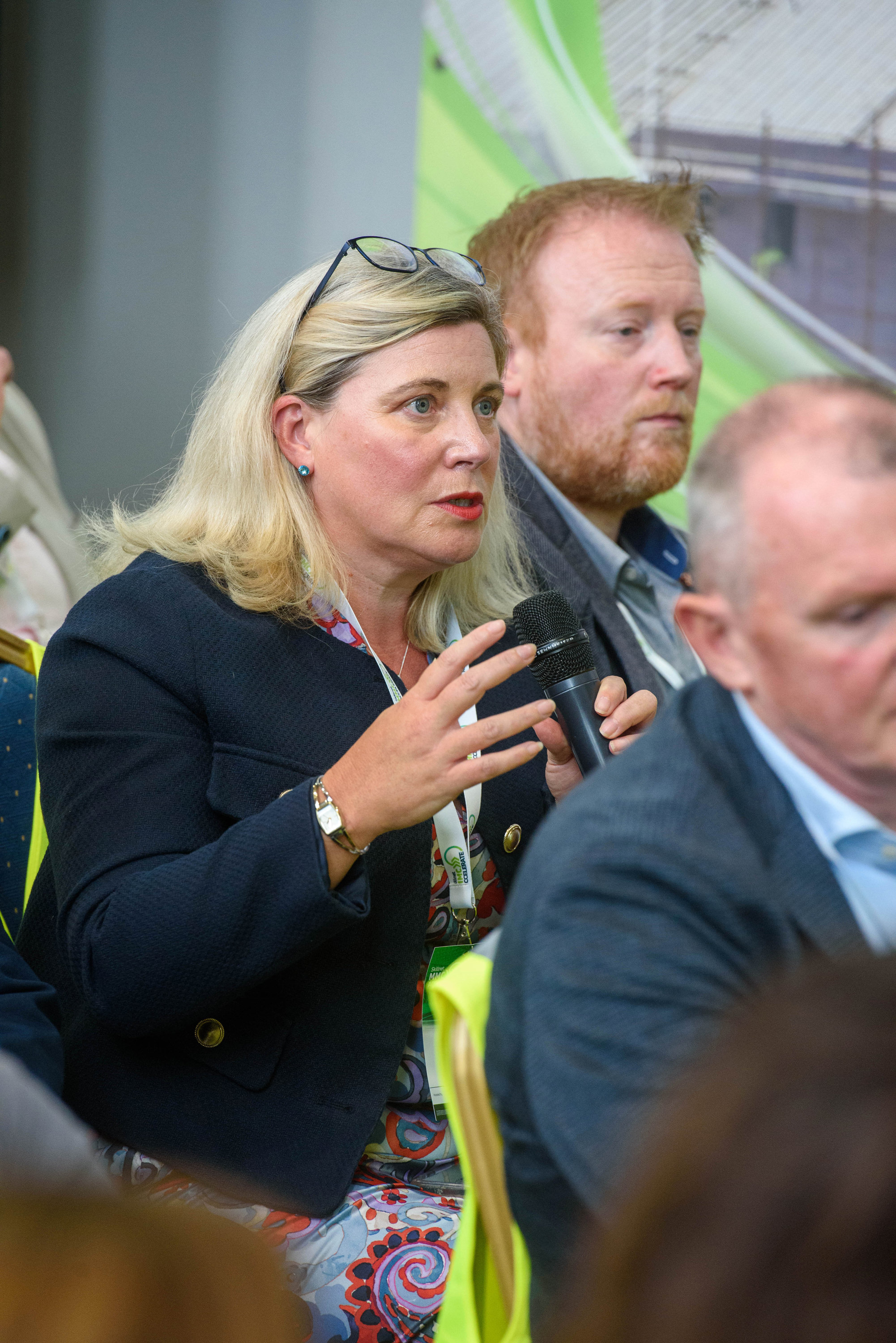 Person in dark jacket holding a camera at an outdoor gathering, wearing a yellow safety vest.