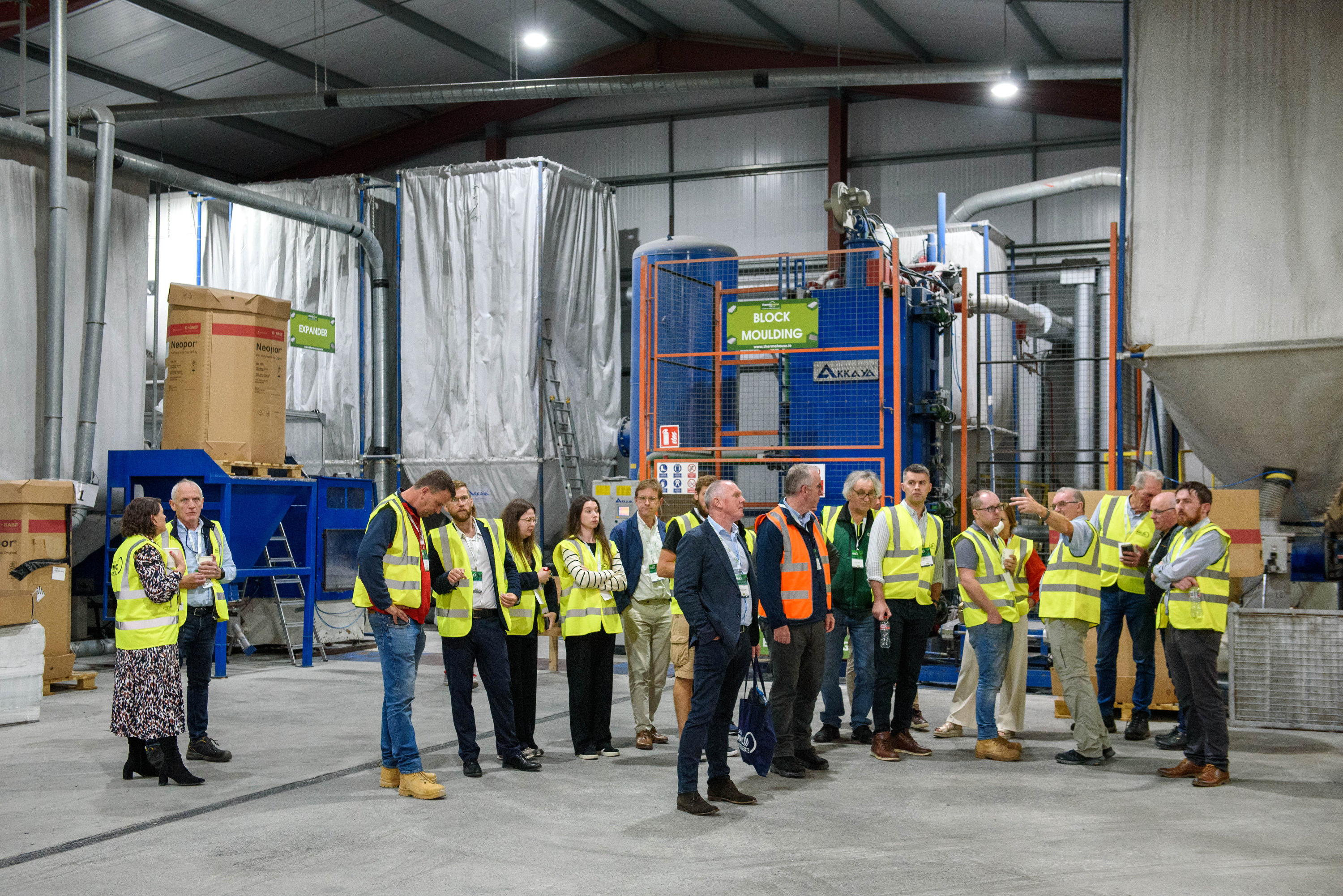 Group of workers in yellow safety vests gathered in an industrial warehouse facility with machinery and equipment.
