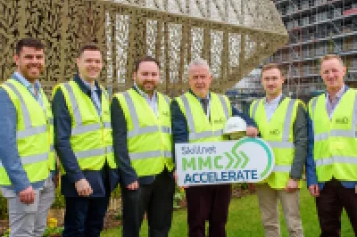 A group of males in high vis vests stand in front of a modern structure, holding a board has the Skillnet MMC Accelerate logo.