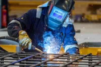 A person in protective clothing and a welding helmet uses a blowtorch to weld steel bars in a manufacturing facility.
