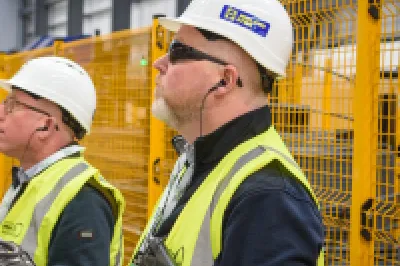 Three people in high-vis vests and hard hats observing machinery inside a construction factory.