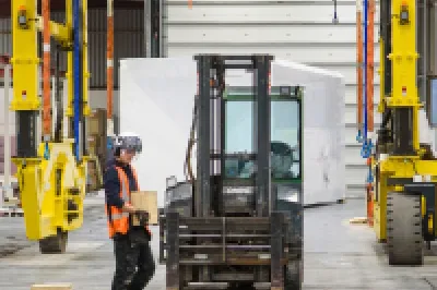 A person wearing a high vis vest and hard hat carries a wooden block through a manufacturing facility passed a forklift.
