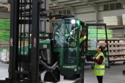 A person in a high vis vest with ear muffs show a tablet screen to a person driving a forklift.