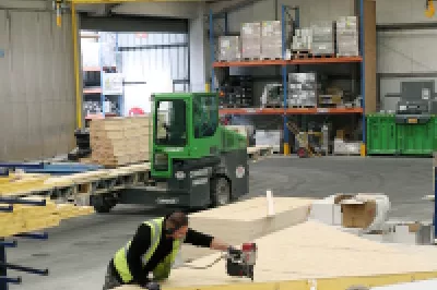 Three people in high vis jackets use power tools to cut wooden frames while two people in high vis jackets with a tablet observe.