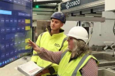 Two people in high-vis vests reviewing data on a large screen, with a 3D printer visible in the background.