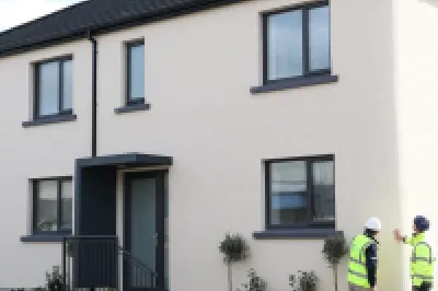Two people in high vis jackets and hard hats inspect modern white semi-detached houses.