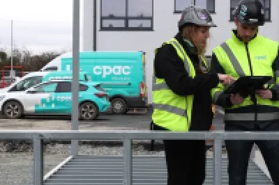 A male and a female read from a tablet inside a prefabricated steel frame set in a car park.