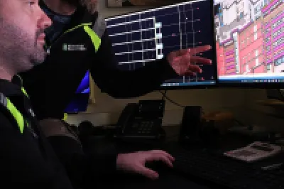 A male points out something in a digital building design on a computer screen to another male.