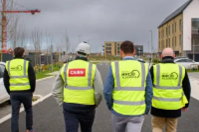 A group of five people in high vis vests walk past a white car through a modern housing development.