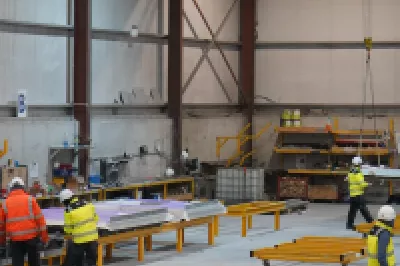 Several people in high vis vests work at machinery stations in a manufacturing facility.