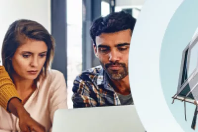 A group of office workers gather around a laptop. To the right, a modular building unit is hoisted by a crane.