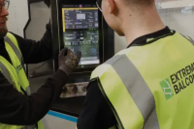 A male with a high vis vest and gloves explains digital readings on a machine screen to another person in a high vis jacket.