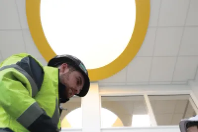 A male in a hard hat with a tablet shows the screen to another male in a hard hat standing on a ladder. 