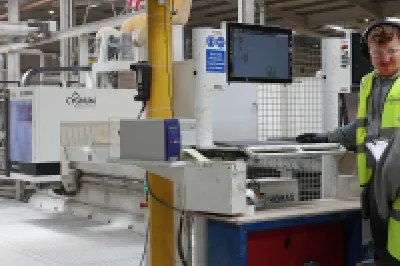 Two workers in high-vis vests monitoring a 3D printer control station inside a large manufacturing facility.