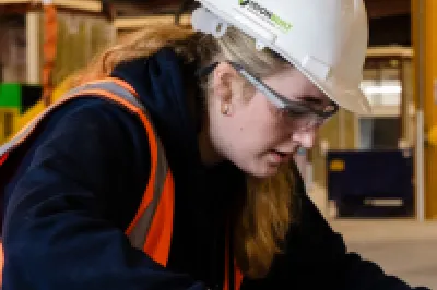 Person in high-vis vest and hard hat inspecting the quality of a metal frame in a factory.