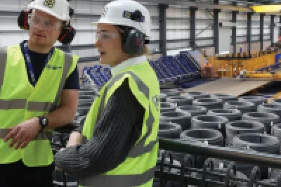 A male with a high vis vest, hard hat, and ear muffs points at something to a female in a high vis vest, hard hat, and ear muffs.