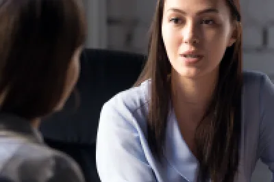 Two females in business clothing sit at a desk in a modern office space talking.
