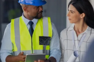 A male in a hard hat and high vis jacket holding a tablet explains something to two people in business clothing.