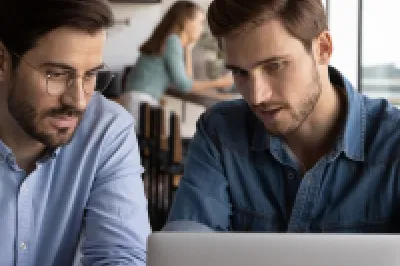 Two males in business shirts sit at a desk and read from a laptop. One takes notes with a pen on a piece of paper.