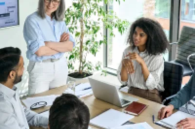 A group of people in business clothing with laptops and pieces of paper in front of them sit around a long table, talking.