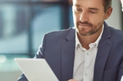 A male in a blue business suit reads from a tablet to another male in a business shirt.