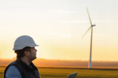 A male in hard hat and vest with an open laptop stands in a field with wind turbines in the background.