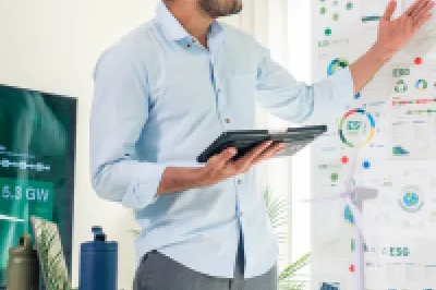 A male in a business shirt stands with a tablet in his hand explaining information on a flip chart to a group of people sat at a table. 