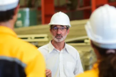 A male with glasses and a hard hat addresses a crowd of people wearing work overalls and hard hats.