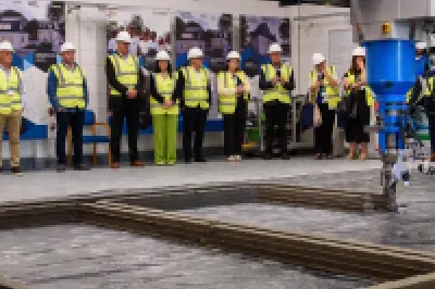 A group of people wearing high-visibility vests and hard hats standing in a row inside an industrial or construction facility, with machinery visible in the foreground.