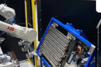 Industrial robotic arms working on components mounted in blue frames inside a manufacturing or training facility. Workbenches and tools are visible in the background.