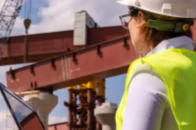 A construction worker wearing a high-visibility vest stands in front of steel structures.