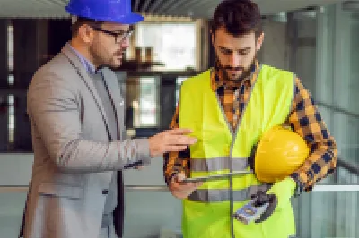Two people wearing hard hats are standing on a construction site, discussing a project.