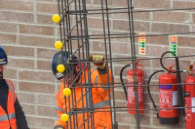 Two construction workers in safety gear standing near a vertical rebar structure inside a building under construction.