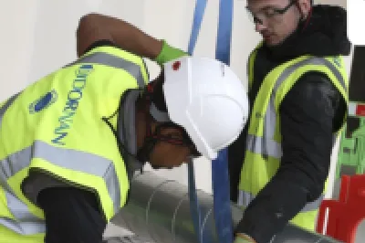 Two construction workers in high vis jackets, white hard hats, and goggles work on tying a pipe