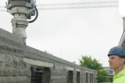 A construction worker in a hard hat and high-vis jacket observes a 3D printer building a concrete wall.
