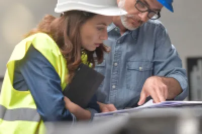Two people wearing hard hats and safety vests reviewing a technical drawing in an industrial setting.
