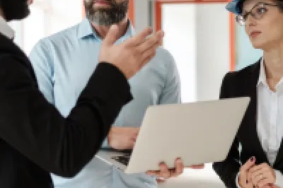 Three engineers wearing formal clothing and safety helmets discussing a construction project indoors, with one holding a laptop.