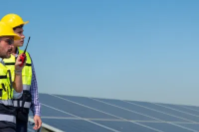 A construction worker wearing a yellow hard hat and safety vest stands beside a solar panel array, holding a toolbox in a sunny outdoor environment.