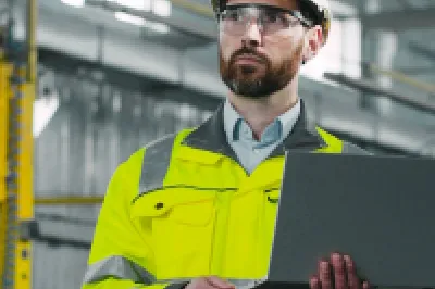 Person in a hard hat and high-visibility jacket using a laptop at an industrial facility.