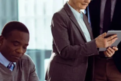 Group of professionals in a modern office having a discussion around a table, with one person showing a tablet to a colleague.