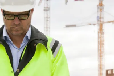 A person in a high-visibility jacket and white hard hat using a tablet on a construction site, with cranes and buildings in the background at sunset.