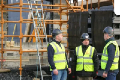 Three construction workers in helmets and safety vests discussing at a building site with scaffolding