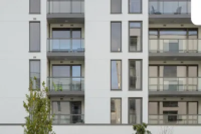 Front view of a modern apartment building with several balconies, large windows, and a landscaped entrance area with plants and trees.