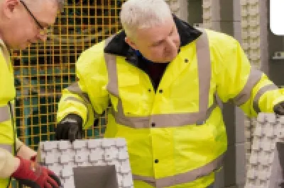 Two people in high-visibility jackets inspecting large grey insulation blocks inside a manufacturing facility, with machinery and safety fencing in the background.